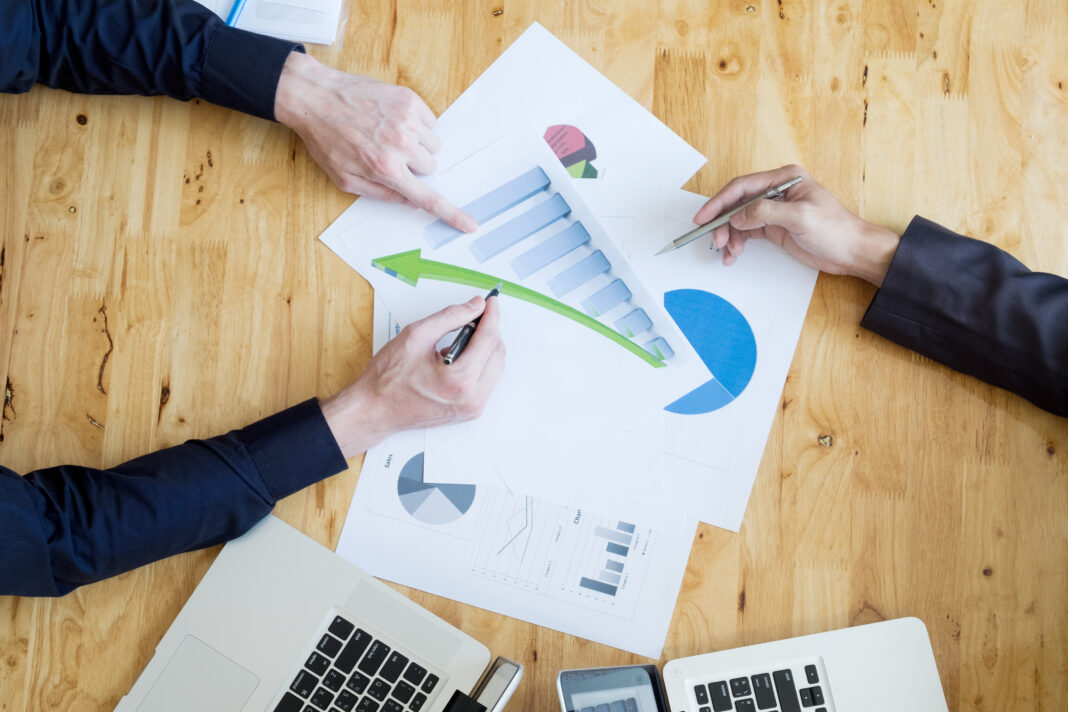 Two business professionals analyzing financial graphs and charts during a meeting. They are pointing to upward trends on the documents laid out on the wooden table, with laptops and tablets in view