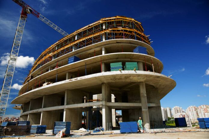 A modern multi-storey concrete building under construction in DHA Lahore, with scaffolding, cranes, and construction materials visible at the site.